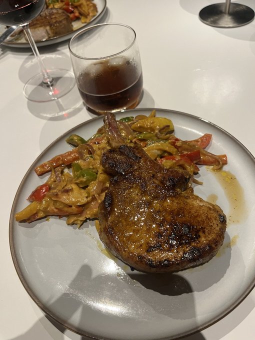 A bone-in pork chop with a dark glaze sits on a white plate alongside sautéed peppers and onions, with a glass of red wine in the background.