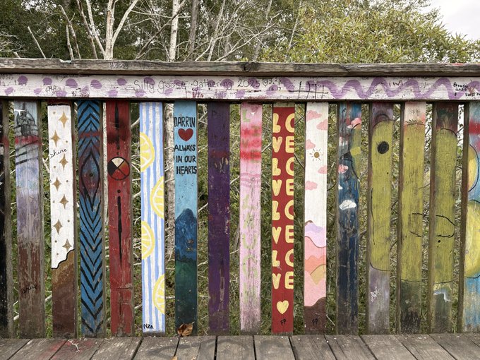 A wooden fence with each plank painted in unique designs and messages, including phrases like 'DARRIN ALWAYS IN OUR HEARTS' and 'LOVE'.