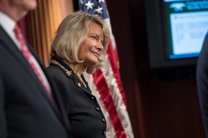 A blonde woman in a dark blazer smiles while standing in front of an American flag, appearing at what looks like an official government event. The image appears to depict Senator Cynthia Lummis in relation to her remarks about the CLARITY Act and crypto sanctions against North Korea's Lazarus Group.