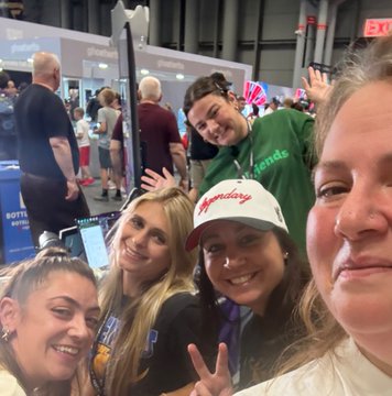 A group of four smiling people pose for a selfie at what appears to be a convention or expo, with one person wearing a 'Monday' trucker hat and another in a green hoodie making peace signs.
