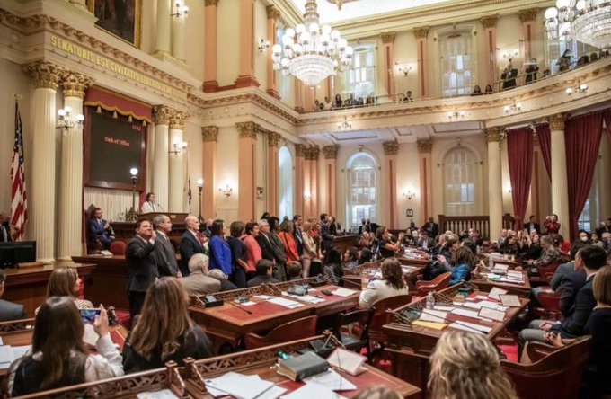 Interior of a grand government chamber with ornate chandeliers, classical architecture, and people seated at desks while some stand near the front, with 'SENATORS REPRESENT' partially visible on the wall.