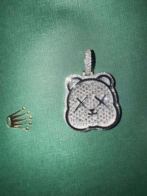 A diamond-encrusted bear-shaped pendant with an 'X' eyes design (the KAWS companion logo) rests on a green fabric surface next to a matching crown-shaped pin.