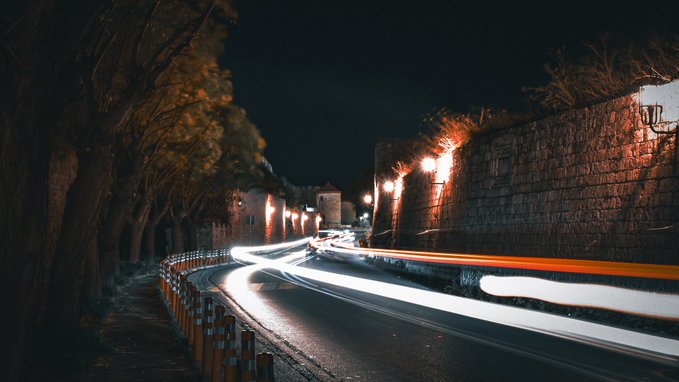 A nighttime long-exposure photograph of a winding road alongside a stone wall, showing streaks of car lights, illuminated street lamps, and a tower in the distance.