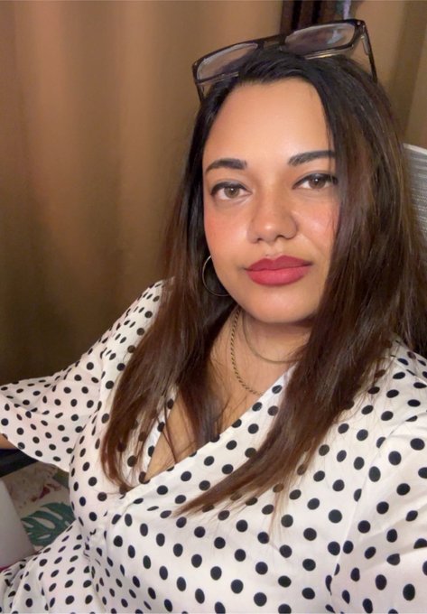 A woman with long brown hair, wearing a white polka dot blouse and hoop earrings, poses indoors with sunglasses perched on her head.