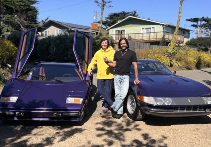 urns of gold and purple, two men pose with cars on a gravel driveway, backed by a house and trees.