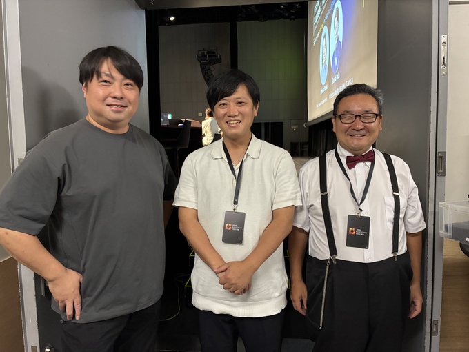 Three men pose together at what appears to be a professional event, with the two on the right wearing lanyards and the man on the far right sporting a bow tie and suspenders. The joking caption about "Ooki-san" likely refers to one of the smiling men, humorously commenting on weight fluctuations compared to Bitcoin's volatility.