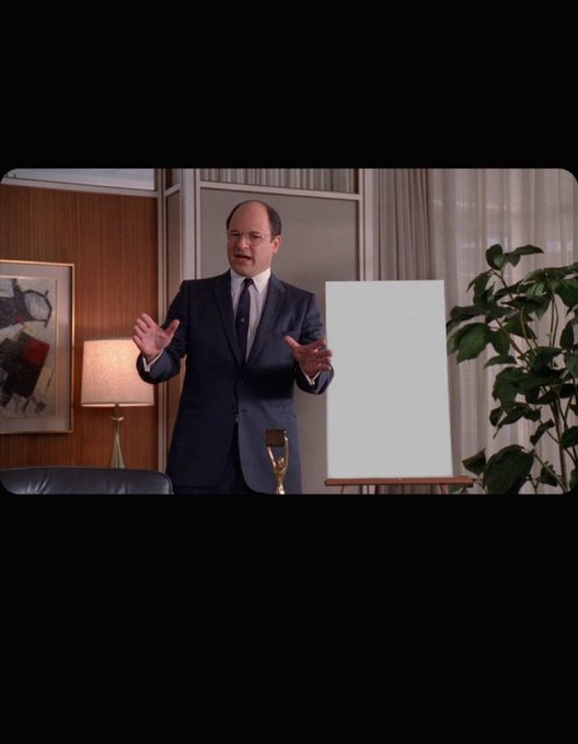 A cinematic still of a balding man in a blue suit gesturing with open hands in a mid-century modern office setting, with a lamp, framed art, a white canvas on an easel, and a potted plant visible.