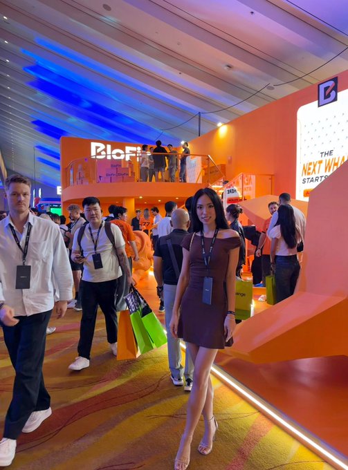 A woman in a brown dress and lanyard poses confidently in the foreground at a vibrant orange event space featuring a "BloFin" booth and a sign reading "THE NEXT WH..." at what appears to be Token2049, completely overshadowing the unsuspecting attendees walking behind her in the candid shot.