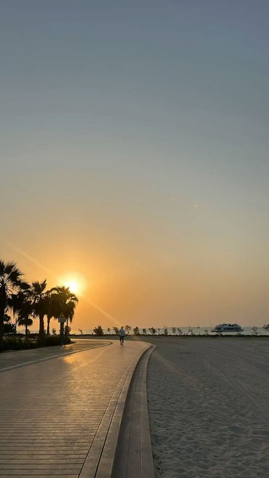 A lone figure walks along a sunlit beach boardwalk at sunrise, with palm trees silhouetted against the golden sky as they greet the morning.
