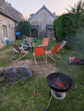 The image shows a peaceful backyard scene with orange deck chairs, a charcoal grill with burgers cooking, and a charming stone cottage in the background. It relates to the post by illustrating the relaxed European vacation lifestyle made possible through Web3 earnings, capturing a tranquil evening of outdoor dining during the speaker's two-month stay abroad.