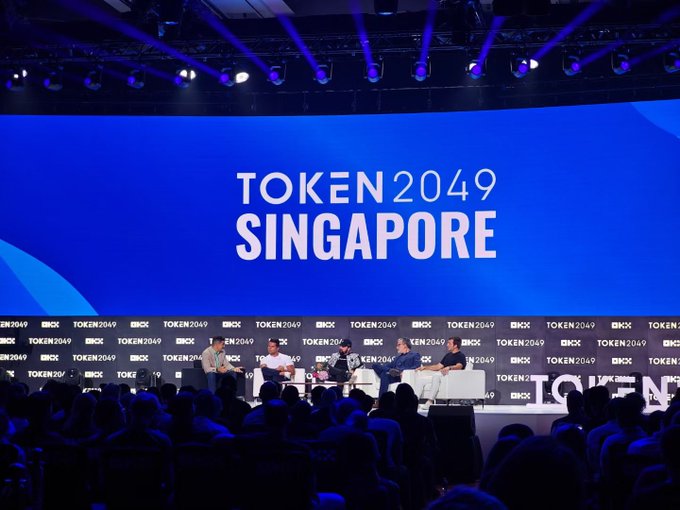 A stage at the TOKEN2049 Singapore conference with the event logo displayed on a large blue screen, where several panelists are seated in front of an audience.