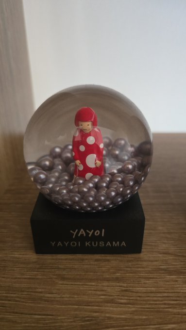 A decorative glass snow globe containing a small figure in a red polka-dot hooded outfit standing among purple metallic beads, mounted on a black base labeled 'YAYOI KUSAMA'.