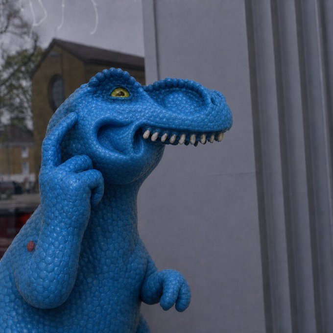 A blue plastic dinosaur toy figure (resembling a raptor) with its hand raised to its face stands outside near a building and street.