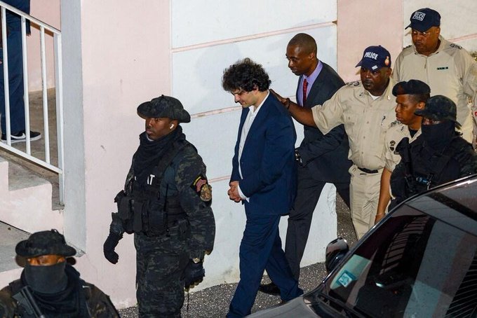 A news photograph showing a young man with curly hair in a blue suit being escorted by police officers and armed personnel in tactical gear, apparently outside a building.