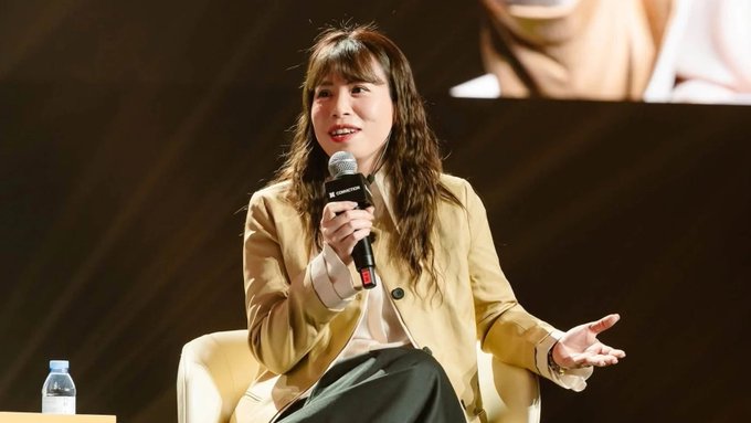 A woman with bangs wearing a tan blazer speaks into a microphone labeled 'Samsung' while seated on stage.