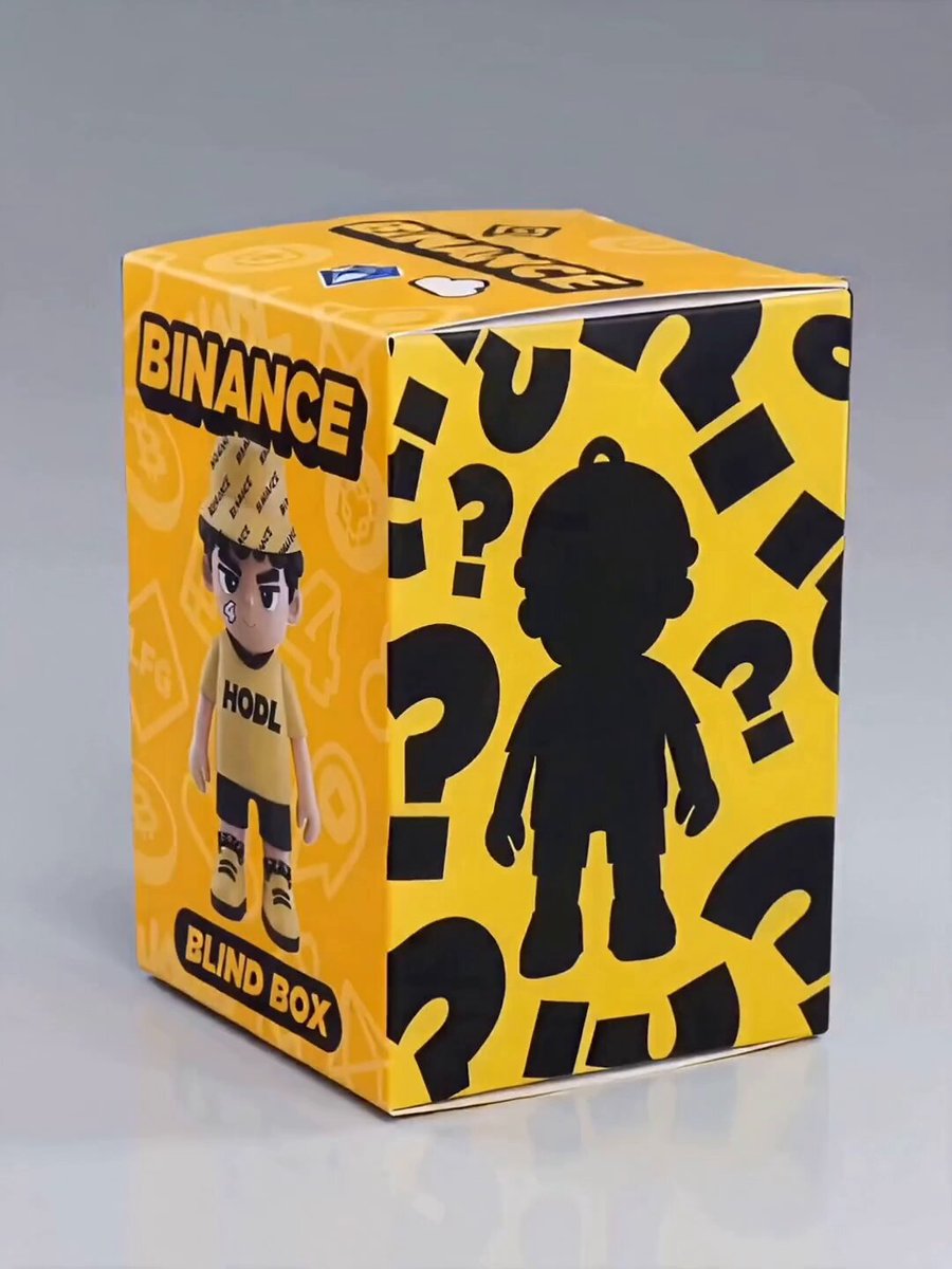 The image features a yellow box with black text that reads "Binance Blind Box" and "Binance Children's Day Gift". The box contains a toy figure of a boy wearing a hat, which appears to be a Hoolish character from the Binance Peripherals series. The toy is sitting on top of the box against a gray background.
