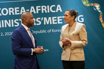 The image shows two people standing next to each other, engaged in conversation. One person is wearing a suit and tie, while the other has a tan blazer over their black pants. They are both facing each other with hands clasped together, suggesting they might be discussing something important or sharing an interesting story. The background features a blue banner that reads "Korea-Africa Ministers' Meeting", indicating this scene takes place at an event related to international relations and cooperation between the two countries.