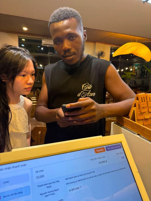 Two people at a café checkout screen, with one wearing a black 'Côte d'Azur' sleeveless shirt looking at a phone, while the display shows an order total of 25,000 ₫ in Vietnamese.