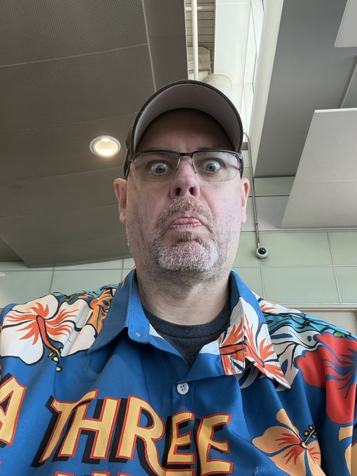 A man wearing glasses, a baseball cap, and a vibrant blue Hawaiian shirt with orange floral patterns makes a wide-eyed, slightly comically exaggerated expression at Houston's Hobby Airport while waiting to depart for the MWBTCSummit. His shirt features the partial word "THREE" in bold yellow lettering across the front, adding to the playful travel-day vibe of the post.