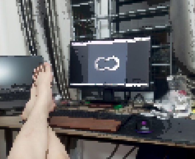 A pixelated image showing a person's bare feet propped up in front of a desk with a laptop displaying a cloud sync icon, alongside a keyboard, mouse, and water bottle.