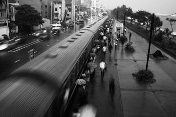 The image shows a train passing by, with people walking along the sidewalk next to the tracks. There are several individuals holding umbrellas as they walk down the street. The scene appears to be captured in black and white, giving it an artistic and timeless quality. The focus on the pedestrians and their activities highlights the everyday life of city dwellers going about their daily routines despite the rainy weather conditions.