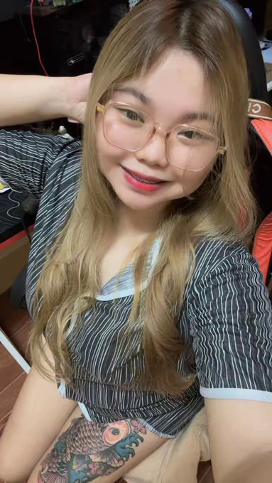 A young woman with long highlighted hair and round glasses smiles at the camera, wearing a striped shirt and sitting in what appears to be a gaming chair, with a colorful fish tattoo visible on her thigh.