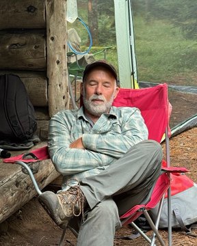 An older bearded man in a plaid shirt and cap sits relaxing in a red folding chair outside a log cabin.