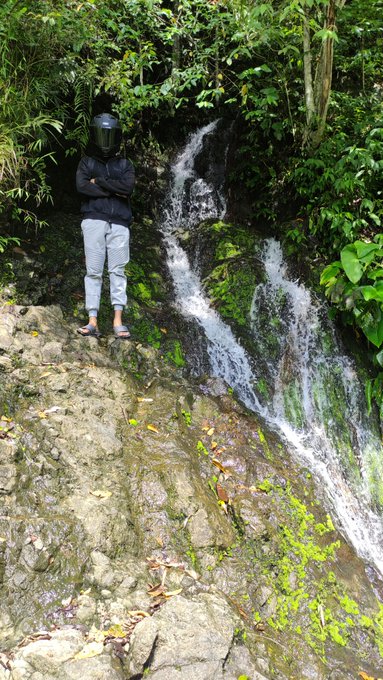A person wearing a black hoodie, gray pants, and a face shield stands with crossed arms on a rocky outcrop beside a small cascading waterfall surrounded by lush green jungle foliage, capturing a quiet moment of perseverance that aligns with the post's motivational message to keep trying and lock in for the new week.