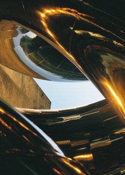 An abstract artistic image showing flowing, ribbon-like metallic shapes with golden highlights curving around an opening that reveals a blue sky.