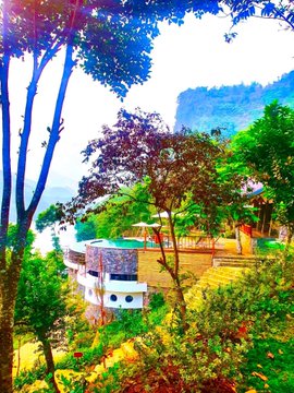 A vibrant scene showing a modern white building with a pool nestled among colorful trees and cliffs, viewed through foliage.