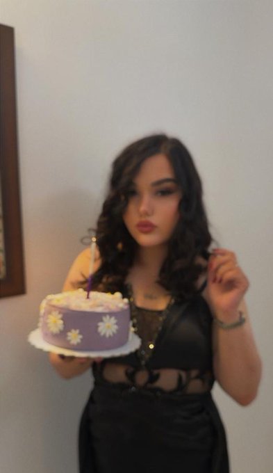 A young woman with long dark hair and bold makeup poses while holding a small purple and white daisy-decorated birthday cake with a single lit candle, celebrating her milestone 25th birthday as referenced in the caption "Dear 25." She wears a stylish black cutout dress and appears ready to make a wish, capturing an intimate birthday moment.