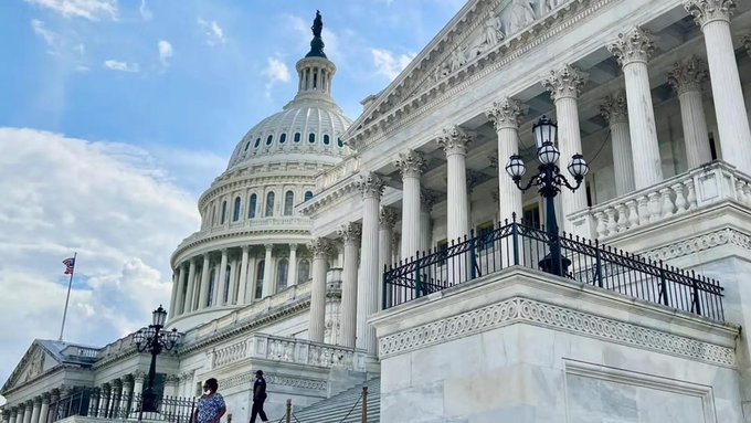 A daytime photograph of the United States Capitol building, featuring its iconic dome and columns, with two figures walking up the steps and an American flag flying.