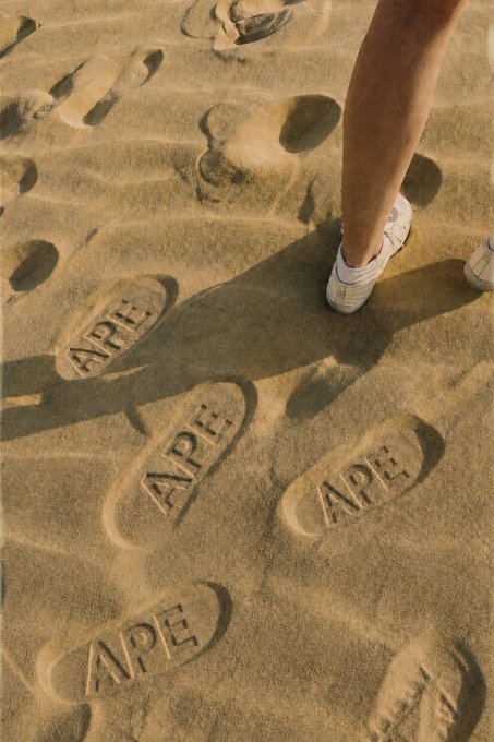 A close-up of a person's legs walking on sandy beach, with footprints in the sand spelling out the word 'APE' repeated multiple times.