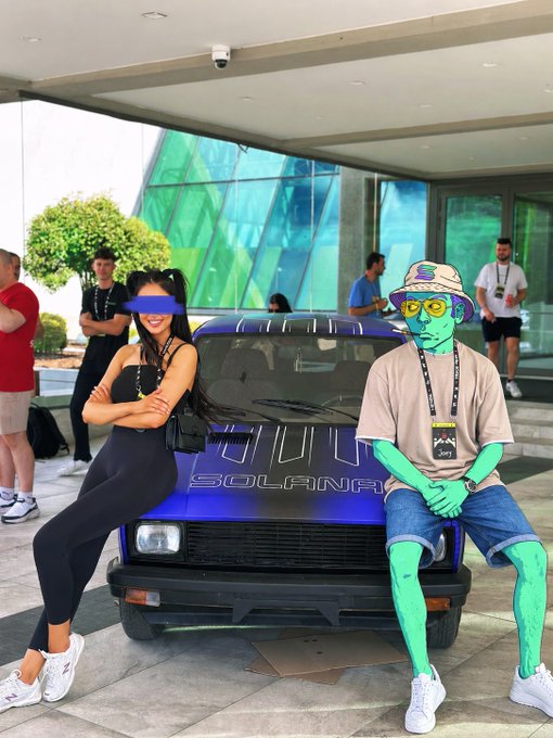A photo of a woman and a person painted entirely green posing in front of a dark blue car with 'SOLANA' written on the front.