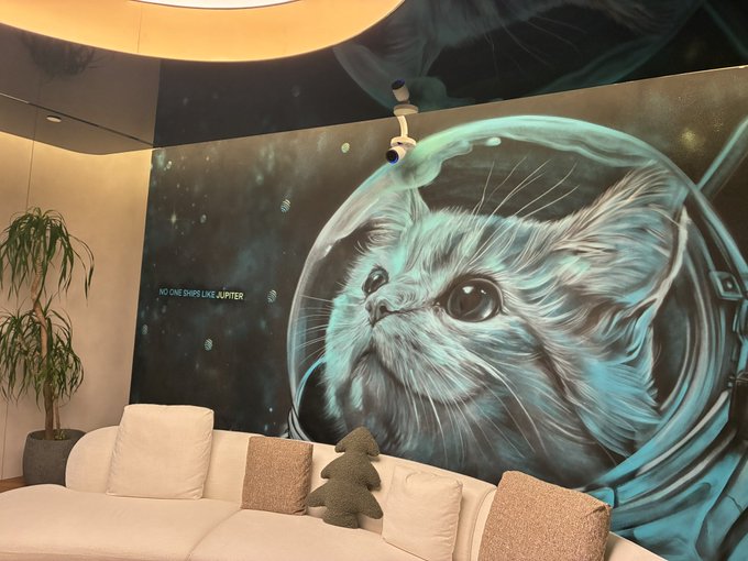 A white couch with pillows sits beneath a striking space-themed mural featuring a cat wearing an astronaut helmet, accompanied by the text "NO ONE OWNS LIKE JUPITER"—the JupiterExchange brand slogan—on the wall. The cozy lounge setting with the playful cosmic cat artwork reflects the hospitality of Jupiter's Kuala Lumpur location, likely a lounge or office space.