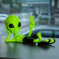 A 3D rendering of a green alien figurine lounging and making a peace sign with its hand.