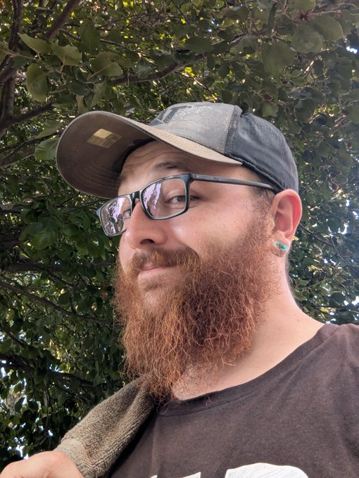 A bearded man wearing glasses, a trucker hat, and blue ear plugs smiles for a selfie against a backdrop of green foliage.