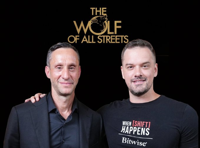 Two men pose together in front of a "The Wolf of All Streets" backdrop, with the man on the right wearing a Bitwise t-shirt that reads "When Shift Happens," reflecting the podcast episode featuring Scott Melker, host of The Wolf of All Streets, alongside fellow crypto host Kevin.