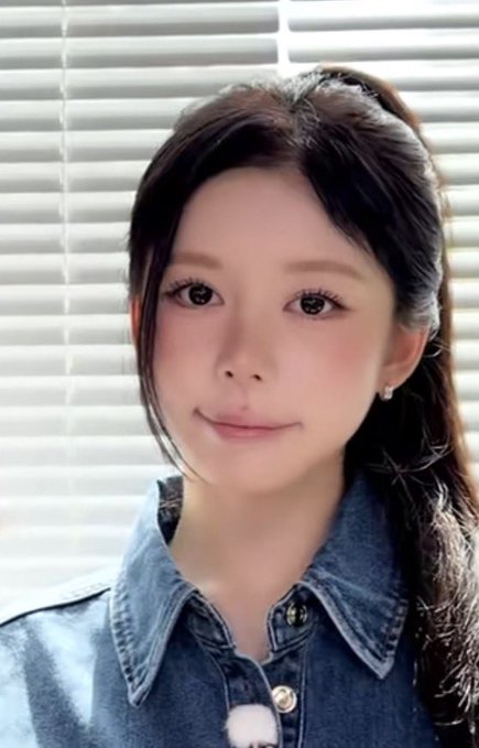 Selfie of a young woman with dark hair wearing a denim jacket, with sunlight streaming through window blinds behind her.