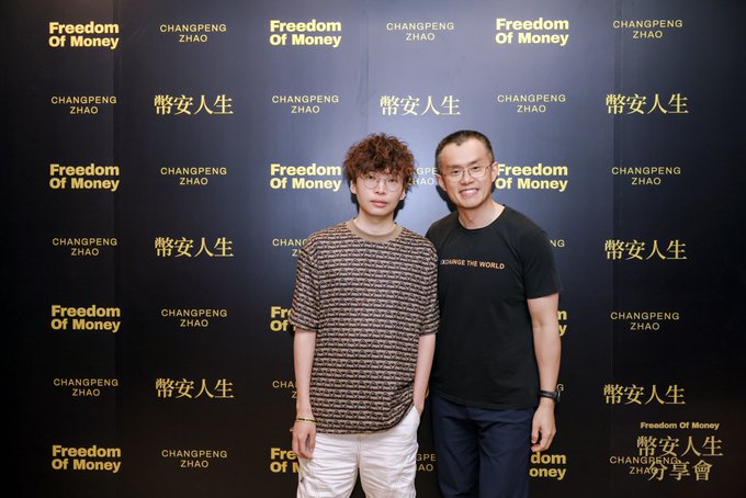 Two men pose in front of a backdrop featuring "Freedom Of Money," "CHANGPENG ZHAO," and "幣安人生" (Binance Life) branding, with one wearing a "CHANGE THE WORLD" t-shirt, capturing a meeting between Binance founder CZ and "Teacher Tony" in Hong Kong to discuss the platform's diversified U.S. stock operations.