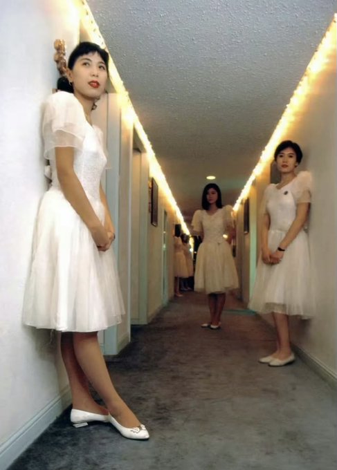 Three women wearing identical white vintage-style dresses standing in a hallway decorated with string lights.
