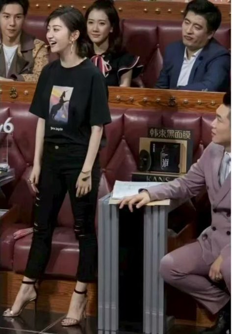 The image shows a woman standing on a TV set wearing a black graphic t-shirt and ripped black jeans with heeled sandals, while a man in a pink suit sits beside her, with other panelists in the background. Visible text includes "韩束黑面膜" (Kans Black Mask) on a product display. This connects to the caption about Zhang Xuefeng, a Chinese education expert known for his appearance on talk shows, being asked about his views on the actress Jing Tian, whose elegant presence contrasts with the casual, smiling subject in the photo.