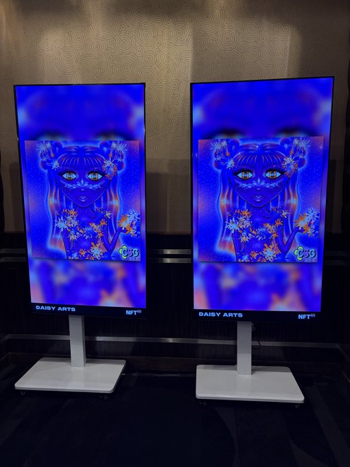 Two illuminated digital displays showcase the same vibrant blue NFT artwork by Daisy Arts, featuring a stylized female figure adorned with floral accents, with "DAISY ARTS NFT" labeled at the bottom of each screen, likely displayed at NFT NYC to celebrate the artist's work.