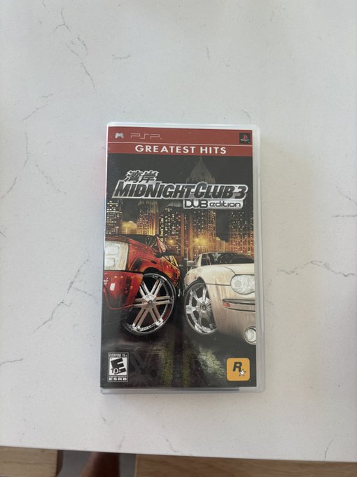 A PSP game case for 'Midnight Club 3: DUB Edition' Greatest Hits edition lies on a white marble surface, featuring two customized cars on the cover.