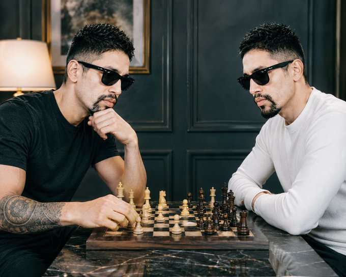 The image shows two men sitting at a table playing chess, with one man wearing sunglasses and holding a wooden chess piece. The other man has a beard and is also wearing sunglasses while looking at his opponent's position. They are both focused on their game, which appears to be an intense match between the two players.