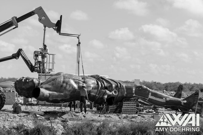 A black-and-white photo shows workers and a crane lifting a massive, humanoid statue in pieces at an outdoor site, with the watermark "AMY DOEHRING" visible in the lower right corner. The scene ties to the "$PROMETHEUS" post by suggesting an Elon Musk-themed sculpture project is being assembled and will soon be unveiled.