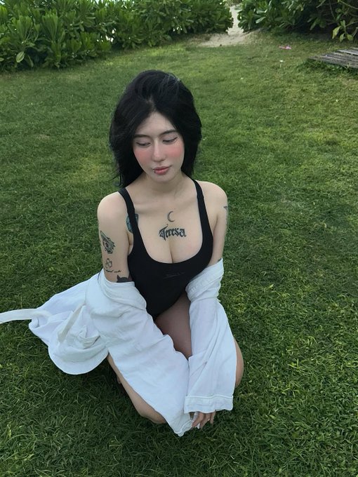 A young woman with dark hair sits on green grass wearing a black bodysuit under a white shirt, displaying a 'Finesse' tattoo with a crescent moon on her chest along with other visible tattoos on her arms.