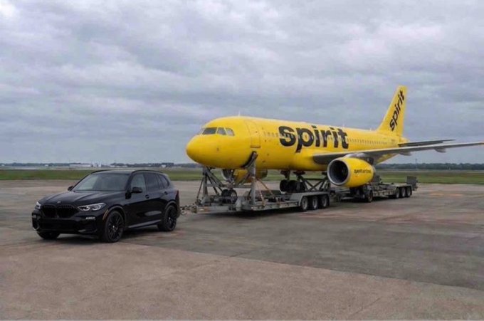 A black BMW SUV tows a large Spirit Airlines yellow airplane on a trailer across an airport tarmac under cloudy skies.