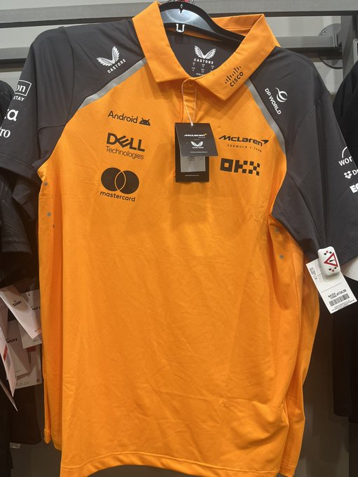 An orange McLaren Formula 1 team polo shirt displays sponsor logos including Android, Dell Technologies, Mastercard, Cisco, McLaren, and OKX, hanging on a rack with Castore branding.