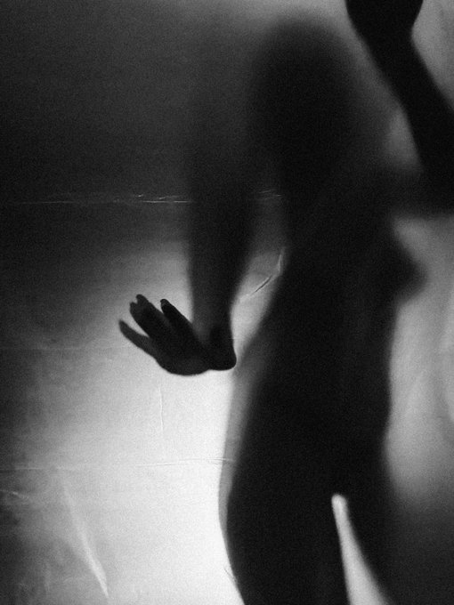 A ghostly female figure presses her hand against a translucent surface in a moody, high-contrast halftone photograph, her shadowy silhouette creating a haunting interplay of light and dark. The image perfectly captures the tension described in the caption—an alluring yet unattainable presence that invites curiosity while simultaneously signaling "don't touch," with the hand's outstretched gesture serving as both a soft welcome and a firm boundary.